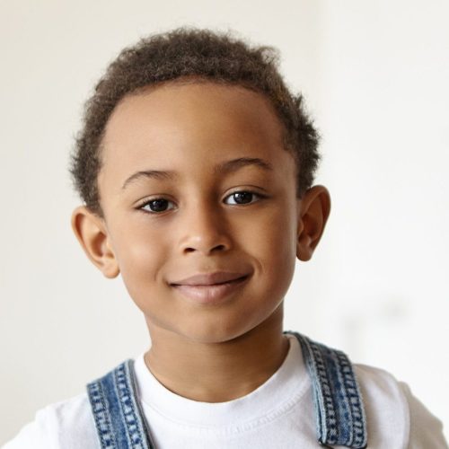 Portrait of handsome cheerful little boy of African origin posing indoors, having confident joyful facial expression, looking at camera with happy smile, dressed in denim jumpsuit over white t-shirt